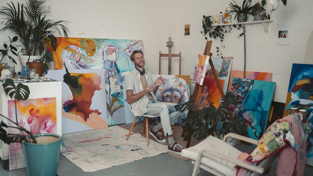 about-img-01 Artist in a vibrant studio surrounded by colorful paintings and lush plants, creating art with joy.